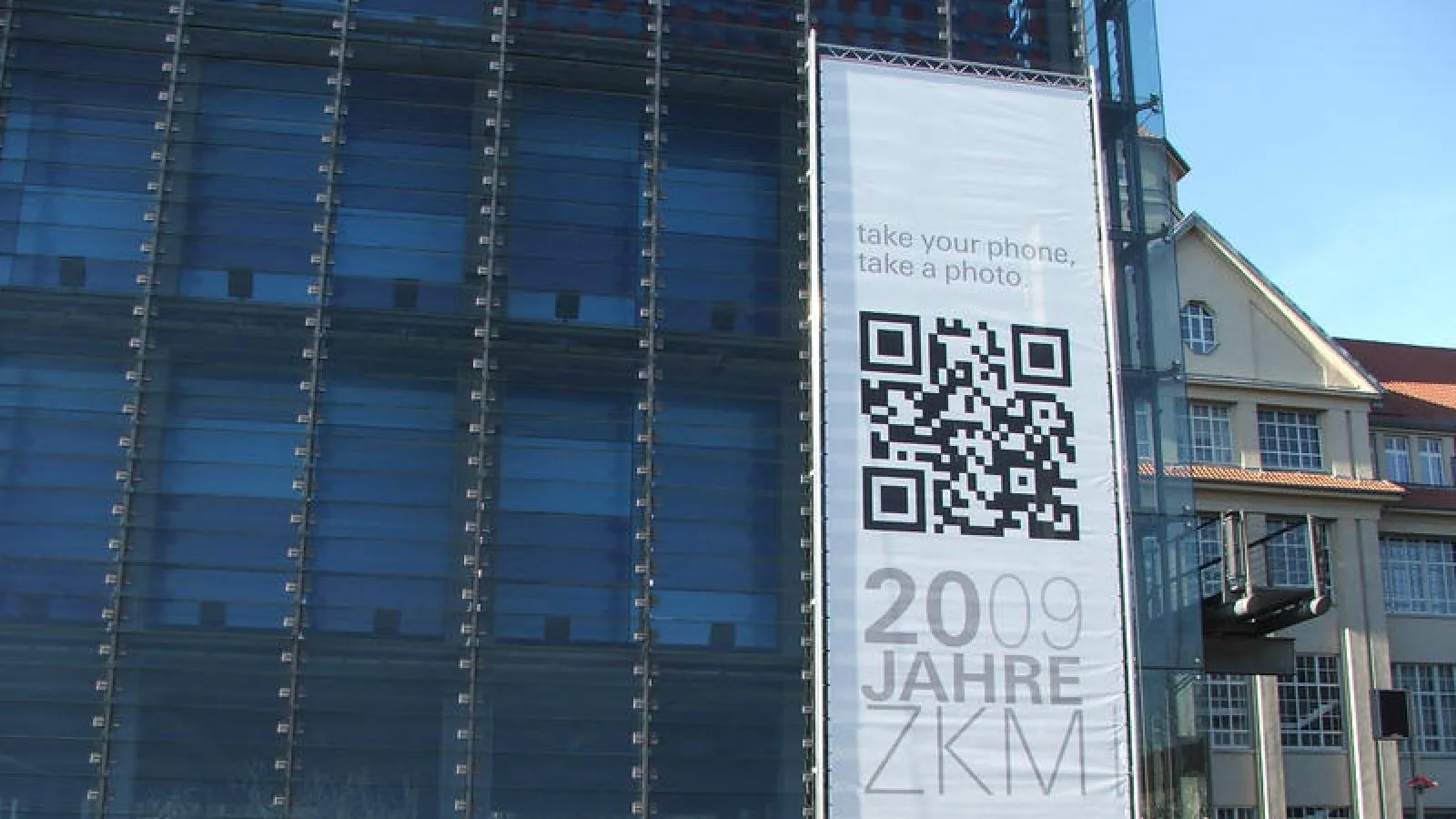 A large banner on the glass facade of the ZKM in Karlsruhe for its 20th anniversary in 2009, showing a QR code, the text take your phone, take a photo, and 2009 Jahre ZKM
